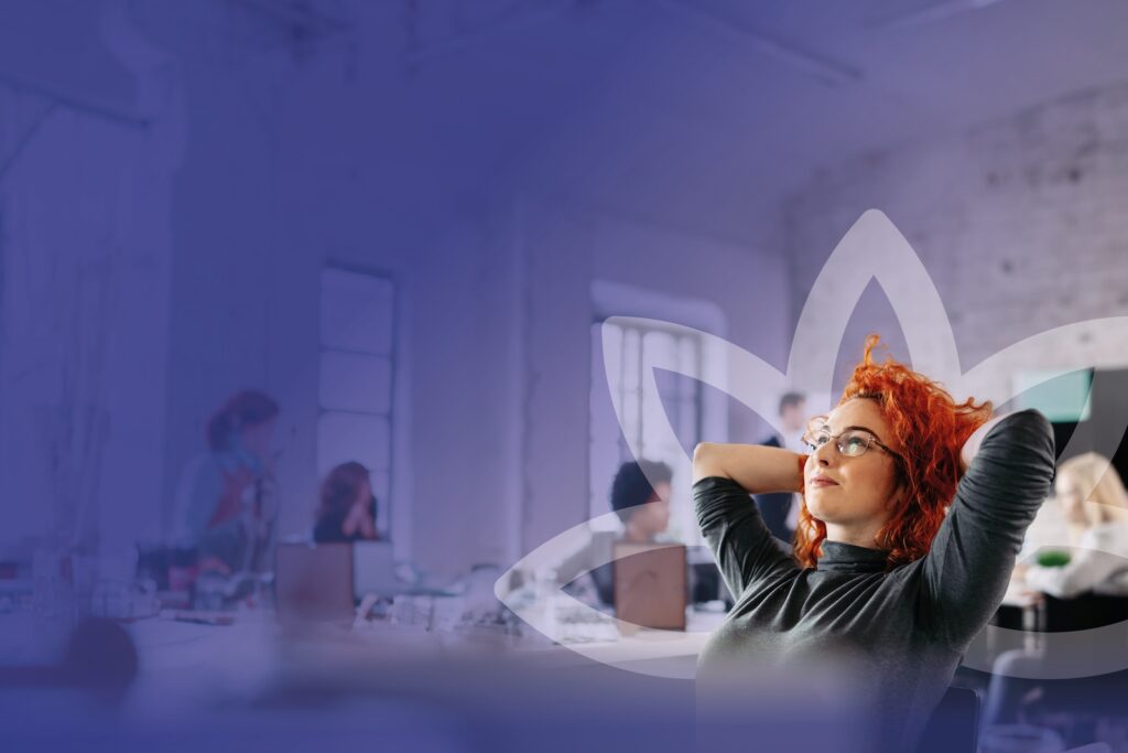 A red-haired professional young woman laying back in her chair with her arms behind her head. She is visibly at peace as she looks off into the distance.