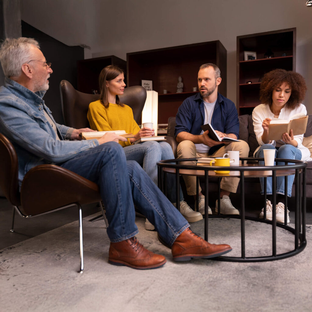 Four adults, sat on chairs around a circle table, reading, talking and drinking tea