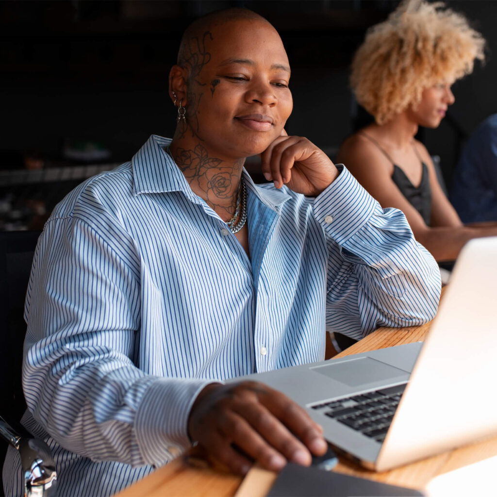 A tattooed woman sat at a desk, smiling and looking at her laptop