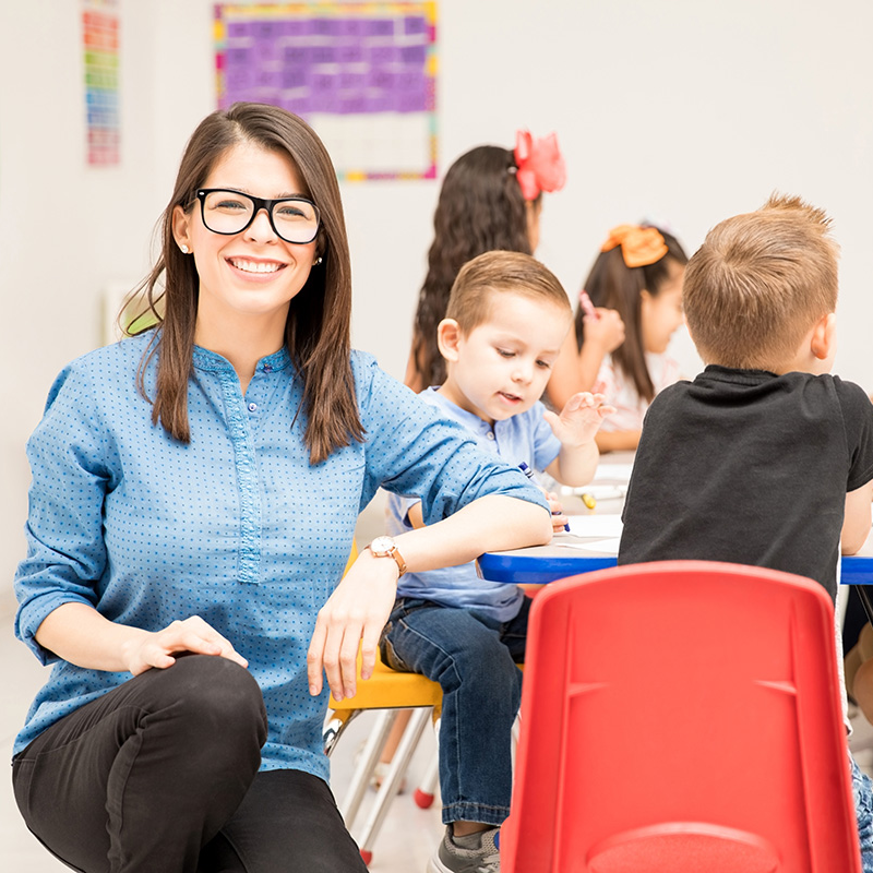 Image of a teacher looking at the camera smiling while her students sit behind her and work.