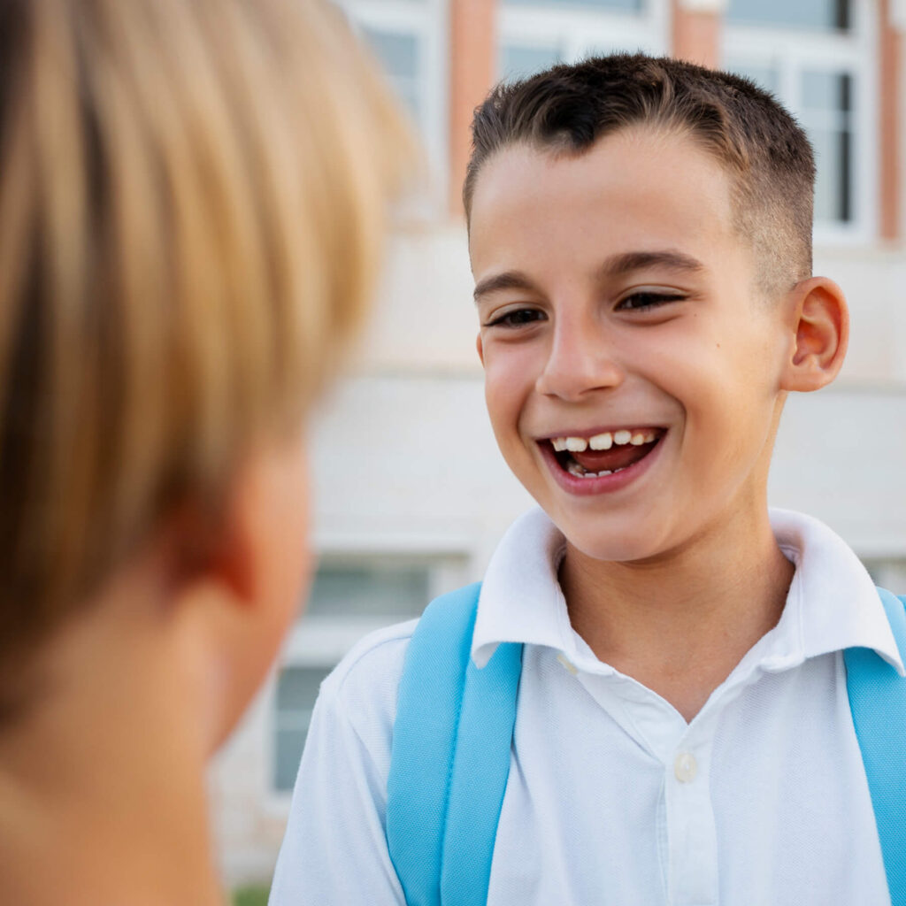 A young boy in his school uniform, wearing a blue backpack, talking and laughing at another student