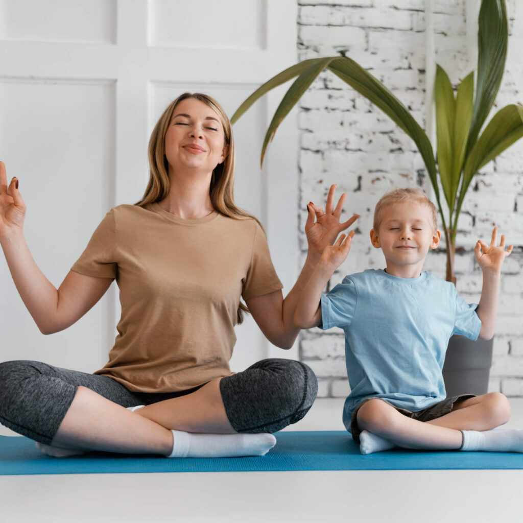A mother and her son sat on a yoga mat, both have their eyes closed and the legs crossed, enjoying and having fun while meditating.