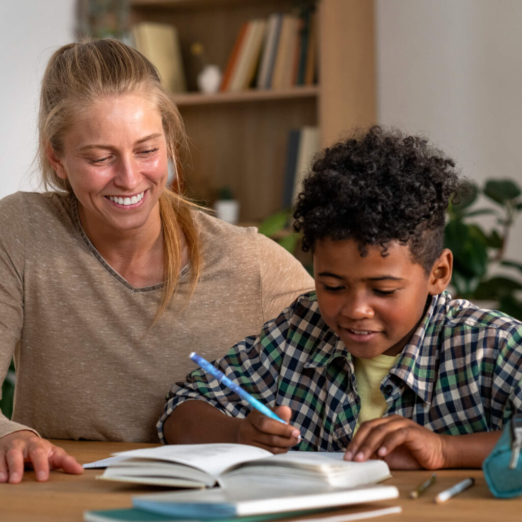 A mother helping her son, smiling, while he is writing in his notebook