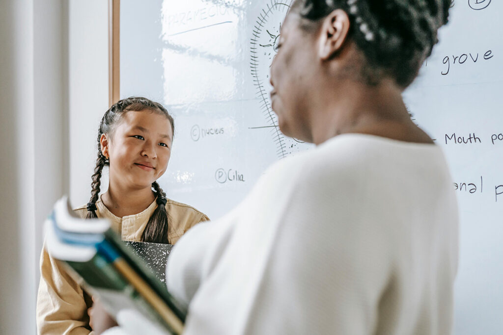 A student looking and smiling at her teacher, while her teacher holds books.