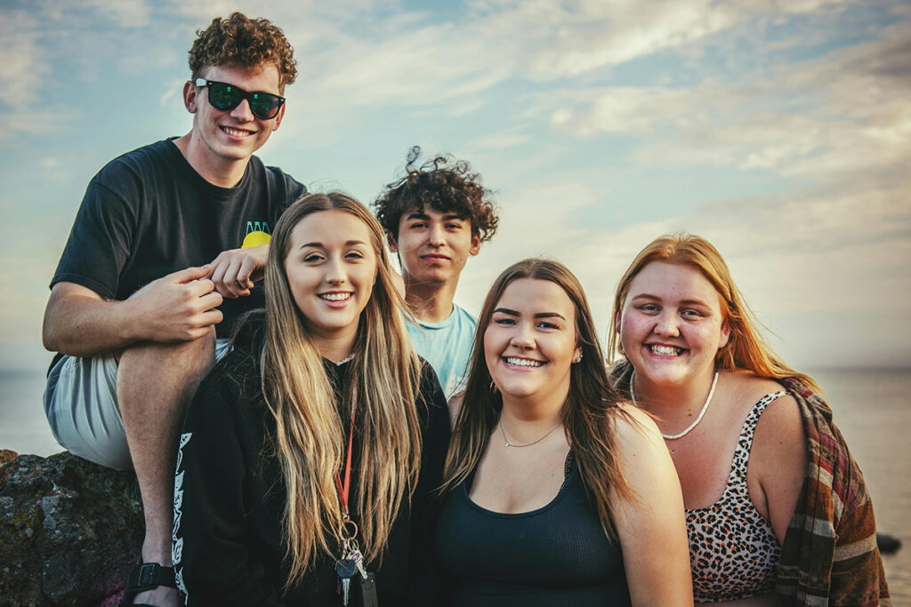 Five teenagers, sat outside at the beach. They're all looking at the camera and smiling.