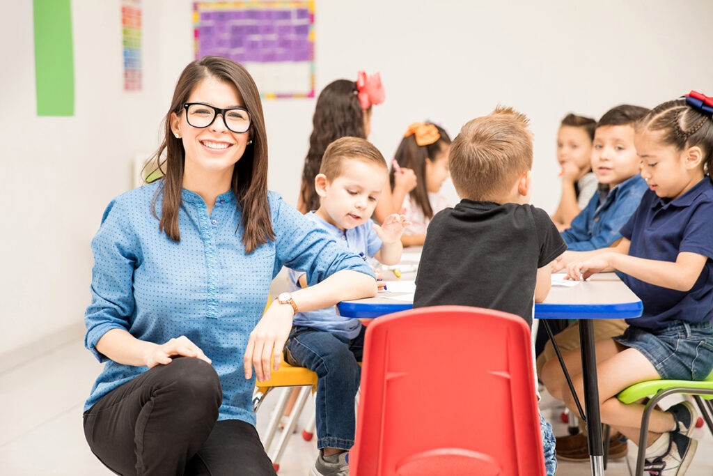 Image of a teacher looking at the camera smiling while her students sit behind her and work.