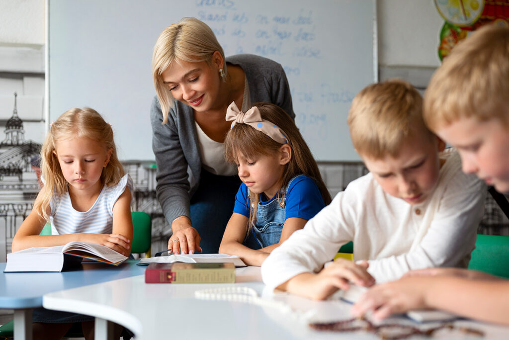 A teacher leaning over her students while her students are studying. The teacher is smiling and helping her read a word.