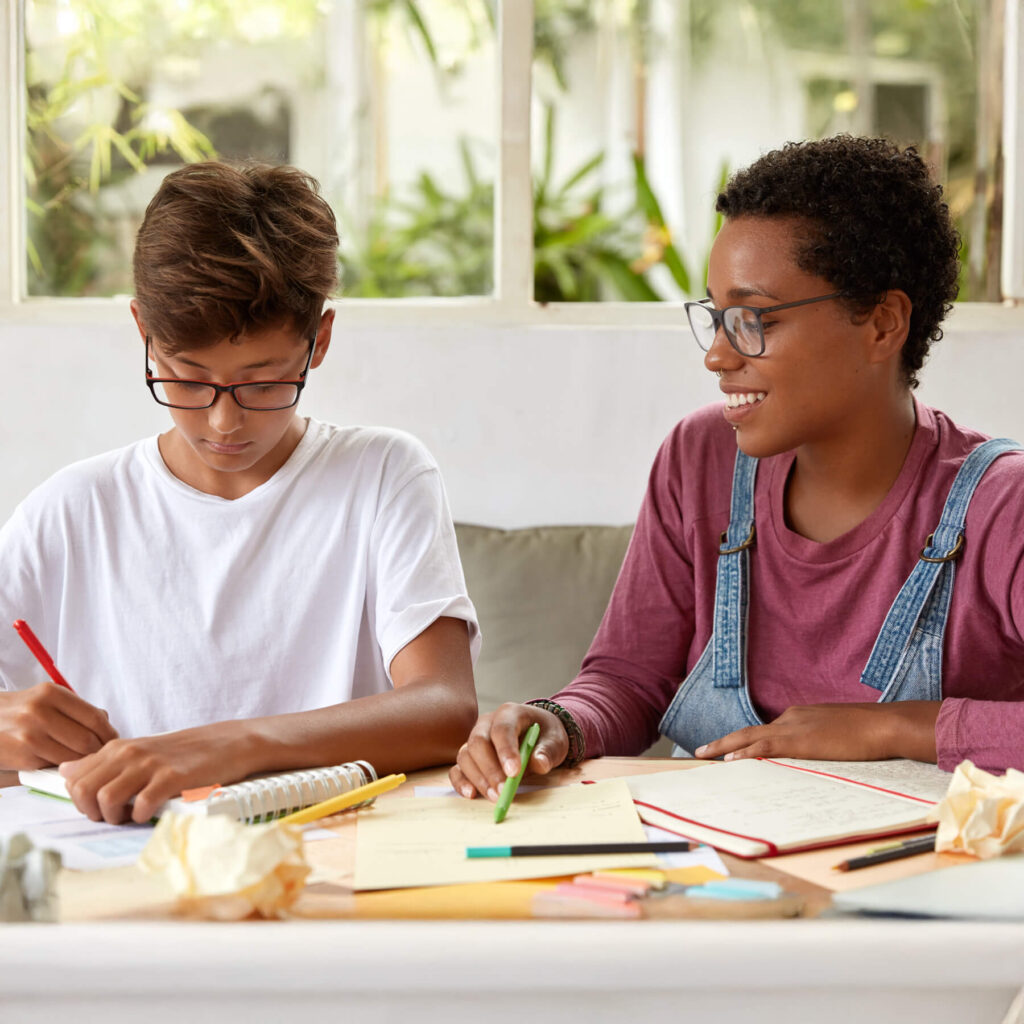 A youth group worker, wearing glasses and smiling, helping a young teenage boy as he writes in his notebook